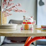 A white and gold lucky cat figurine with pink accents sits on a beige tabletop in a softly blurred indoor setting with pink blossoms and furniture in the background.