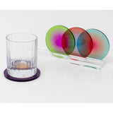A clear tumbler glass on a purple coaster sits beside a transparent acrylic stand holding three overlapping circular colored coasters in green, red, and blue.
