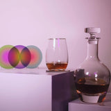 A clear glass decanter and a clear stemless glass with amber liquid, styled on white pedestals against a pale pink-lilac background, with three overlapping iridescent circular coasters on the left.