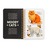 An open spiral-bound sticker book titled 'MOODY CATS' with a black left page containing text and a white right page showing several cat-shaped stickers, including one orange cat, one white cat, and a small orange tabby.