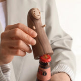 A person holding a wooden bottle opener shaped like the character No-Face from Spirited Away, using it to open a bottle cap.