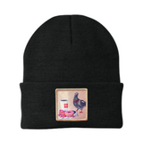 A black knit beanie with a folded cuff featuring a square embroidered patch. The patch depicts a pigeon, a pink donut and a cup of coffee