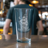 A clear pint glass with an etched sailing ship and crescent moon design, set on a wooden bar counter with a blurred bartender in the background.