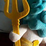 A close-up shot of a plush toy holding a yellow trident, with a teal hair accessory and a white body visible against a dark background.
