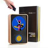 A Klydo Clock with a light wood frame and black face, featuring a circular blue clock face with a stylized figure and a yellow button. A hand is shown holding the black remote control next to the product, with its black and yellow packaging box in the background.