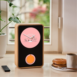 A rectangular smart clock with a light wood frame and a black front panel sits on a tabletop near a window. The clock face is pink with a minimalist cat illustration and white hands, and there is a round orange accent below.