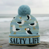 A light blue knit beanie with a pom-pom, featuring a pattern of seagulls and a puffin, with the text 'SALTY LIFE' knitted on the cuff. The hat is photographed outdoors on a rock with a blurred ocean and cloudy sky in the background.