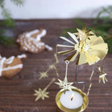 A gold metal tealight candle holder with star and windmill details, displayed on a dark wooden surface with a lit tealight candle, with pine branches and gingerbread cookies in the background.