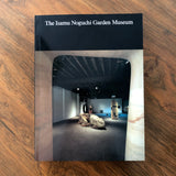 A hardcover book titled 'The Isamu Noguchi Garden Museum' resting on a brown wooden surface. The cover features a black top band with white text and a central photograph of an art gallery interior framed by stone columns.