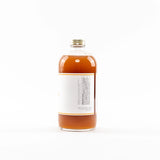 A clear glass bottle filled with amber liquid, featuring a blank white label and a gold screw cap, photographed against a seamless white background.