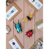 Decorative paper insects on a wooden surface with packaging and tools.