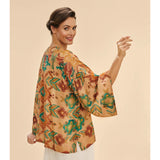 A woman modeling a loose, 3/4 sleeved kimono-style jacket with a warm tan base and an all-over floral and botanical print in rust brown, teal green, and soft orange, photographed against a solid light beige studio background.