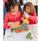 Two young girls are playing with a colorful wooden animal puzzle set on a white table. The children are wearing red long-sleeve shirts, and the wooden puzzle pieces shaped like animals are arranged on the tabletop.