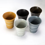 Five ceramic cups in different colors arranged on a white background: mustard yellow with white stripes, black, dark brown speckled, light gray speckled, and slate blue with white stripes.