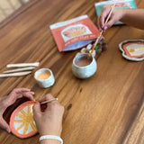 Two people painting clay items on a wooden table with art supplies.