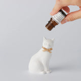 Hand holding a small bottle above a white ceramic cat figurine on a light gray background