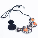 A black cord necklace with circular fabric beads, including two solid black discs and three patterned discs featuring white geometric designs and orange floral motifs, on a white background.