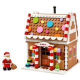 A LEGO-style gingerbread house building set on a white background, with a small Santa minifigure standing beside it. The house has a brown gingerbread facade, white trim, colorful round roof decorations, candy cane pillars, and a front window display.