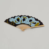 A folding hand fan with a light wood frame, featuring a black background and a stylized illustration of a light blue snake with gold accents.